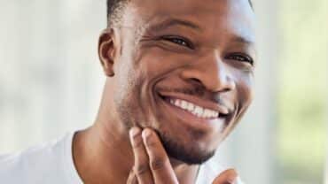 Imagem fechada no rosto de um homem negro. Ele está sorrindo e com a mão direita próximo ao queijo, do lado esquerdo, avaliando possíveis sinais de envelhecimento da pele.