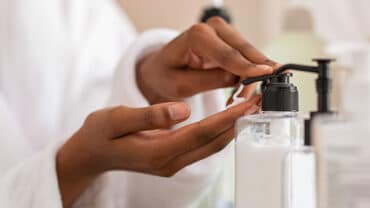 Imagem aproximada de duas mãos em contato com um creme hidratante. Uma está pressionando a válvula, enquanto a outra receber o produto, que pode conter conservantes como parabenos.