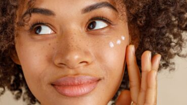 Imagem fechada no rosto de uma mulher. Ela com a expressão levemente sorridente e com os olhos virados para o lado direito. Tem a pele morena, sardas em cima do nariz e está aplicando algumas gotas de creme com os dedos sobre as maças do rosto.