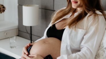 Imagem de uma gestante sentada na cama, com as mãos logo abaixo da barriga. Ela está usando uma calça jeans, uma camisa branca aberta e um top preto. Ela está sorrindo e com os cabelos soltos.