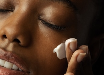 Imagem fechada no rosto de uma mulher, entre a testa e o lábio superior. Ela tem a pele negra e está passando um creme hidratante no rosto, contendo antioxidantes para pele.