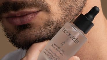 Imagem fechada na região da boca e do queixo de um homem. Ele tem barba e está segurando próximo ao queijo o sérum Natural Light da Noorskin, produto usado para o skincare masculino.