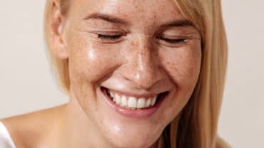 Imagem fechada no rosto de uma mulher. Ela tem a pele e os cabelos claros, sardas, está sorrindo e com os olhos fechados. A pele reflete hidratação, um dos benefícios dos pós-bióticos, prebióticos e probióticos para pele.