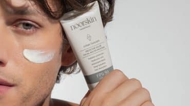 Imagem fechada no rosto de um homem. Ele tem a pele e os olhos claros e os cabelos escuros. Com uma das mãos, segura o Urban Suncare, protetor solar facial da Noorskin, colado ao rosto. Na face está um pouco de protetor solar, demonstrando a importância de proteger a pele todos os dias.