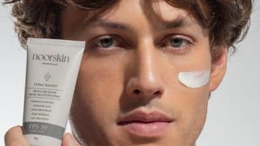 Imagem fechada no rosto de um homem. Ele tem a pele e os olhos claros e os cabelos castanhos. Está segurando o protetor solar Urban Suncare com uma das mãos, colado ao rosto . Tem um pouco de protetor solar sobre a face.