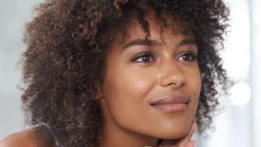 Imagem do rosto de uma mulher. Ela tem a pele negra e os cabelos encaracolados. Está sorrindo levemente e passa a sensação de saúde e bem-estar, como uma pessoa que prática o autocuidado.