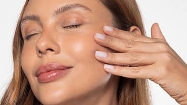 Imagem fechada no rosto de uma mulher. Ela tem a pele e os cabelos claros, está de olhos fechados e passando os dedos sobre as bochechas, um movimento para sentir a textura e a firmeza da pele, benefícios do ativo Skinectura.